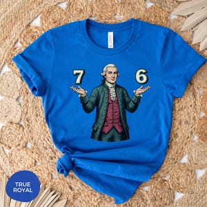 May include: Royal blue t-shirt featuring a graphic of a historical figure holding the numbers 7 and 6. The figure is dressed in a green coat, a red vest, and a white ruffled shirt. The shirt has the text "TRUE ROYAL" on the bottom left.