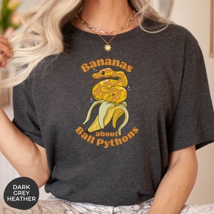 Op de afbeelding: Een donker grijs t-shirt met een afbeelding van een gele banaan met een ball python eromheen gewikkeld. De tekst "Bananas about Ball Pythons" staat op het shirt gedrukt.