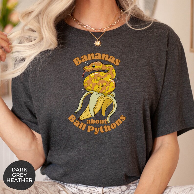 Op de afbeelding: Een donker grijs t-shirt met een afbeelding van een gele banaan met een ball python eromheen gewikkeld. De tekst "Bananas about Ball Pythons" staat op het shirt gedrukt.