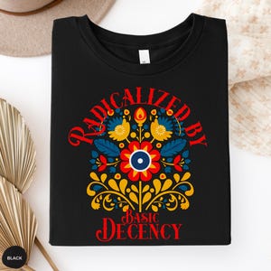 Könnte beinhalten: Schwarzes T-Shirt mit einem farbenfrohen Blumendesign und dem Text "RADICALIZED BY BASIC DECENCY" in Rot. Das Design umfasst blaue, gelbe und rote florale Elemente. Das T-Shirt hat einen Rundhalsausschnitt.