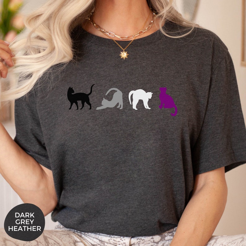 May include: A dark heather grey t-shirt with a row of four black, grey, white, and purple cat silhouettes. The cats are arranged in a line, with the black cat on the left and the purple cat on the right. The shirt is a unisex style.