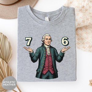 May include: Athletic heather gray t-shirt featuring a cartoon illustration of a man in a historical outfit holding the numbers 7 and 6. The man is wearing a green coat, a red vest, and a white ruffled shirt. The shirt has the words "ATHLETIC HEATHER" printed on it.