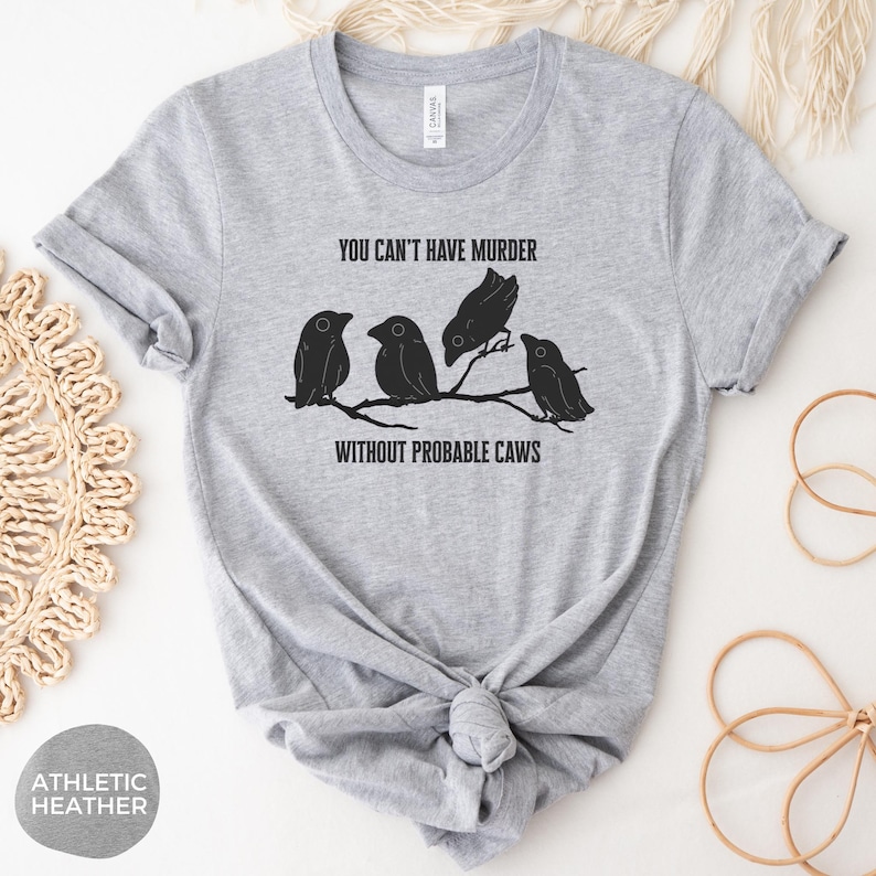 May include: Athletic heather gray t-shirt featuring a black graphic of four birds perched on a branch, with the text "YOU CAN'T HAVE MURDER WITHOUT PROBABLE CAWS". The shirt is knotted at the waist.