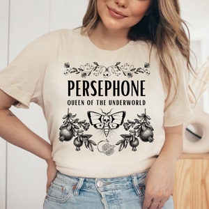 May include: A beige t-shirt with a black graphic design featuring the text "PERSEPHONE QUEEN OF THE UNDERWORLD" with a floral design and a butterfly with a skull.