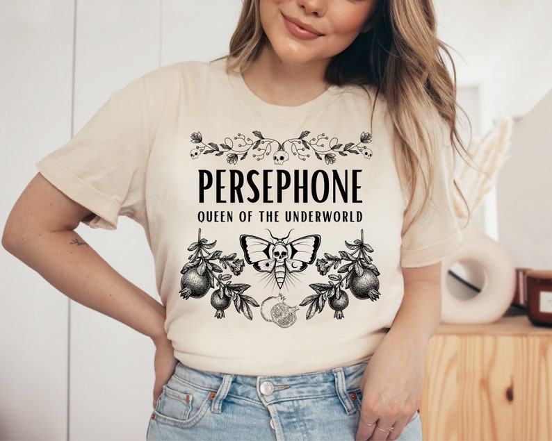 May include: A beige t-shirt with a black graphic design featuring the text "PERSEPHONE QUEEN OF THE UNDERWORLD" with a floral design and a butterfly with a skull.