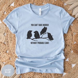 May include: Light blue t-shirt with the text "YOU CAN'T HAVE MURDER WITHOUT PROBABLE CAWS" above a graphic of four black birds perched on a branch. The shirt is made of a soft material.
