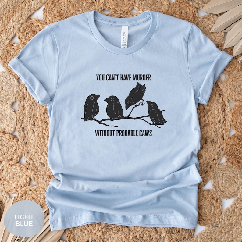 May include: Light blue t-shirt with the text "YOU CAN'T HAVE MURDER WITHOUT PROBABLE CAWS" above a graphic of four black birds perched on a branch. The shirt is made of a soft material.