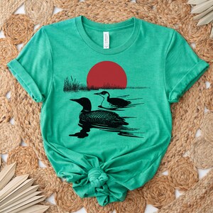 May include: A green t-shirt with a black silhouette of two loons swimming in a lake with a red sun setting in the background.