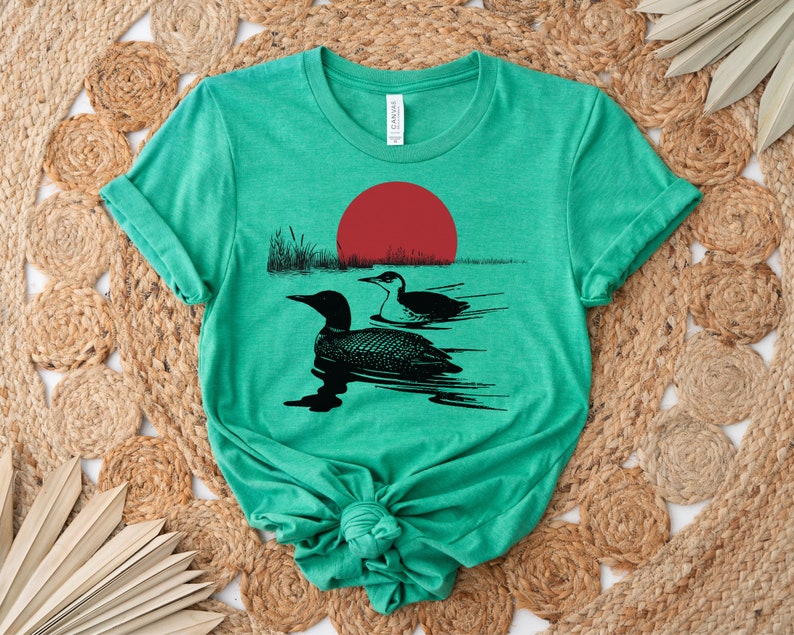 May include: A green t-shirt with a black silhouette of two loons swimming in a lake with a red sun setting in the background.