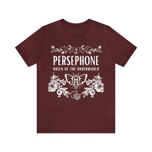 May include: A maroon t-shirt with a white graphic design featuring the text "PERSEPHONE QUEEN OF THE UNDERWORLD" with a moth and pomegranate imagery.