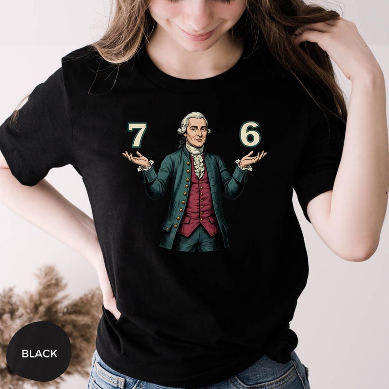 May include: Black t-shirt featuring a graphic of a man in a colonial outfit holding the numbers 7 and 6. The man is wearing a green coat, a red vest, and a white ruffled shirt. The numbers are in a green font.