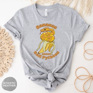 Op de afbeelding: Een grijs T-shirt met een afbeelding van een gele en oranje ball python die om een ​​bos bananen is gewikkeld. De tekst "Bananas about Ball Pythons" is in oranje en geel gedrukt.