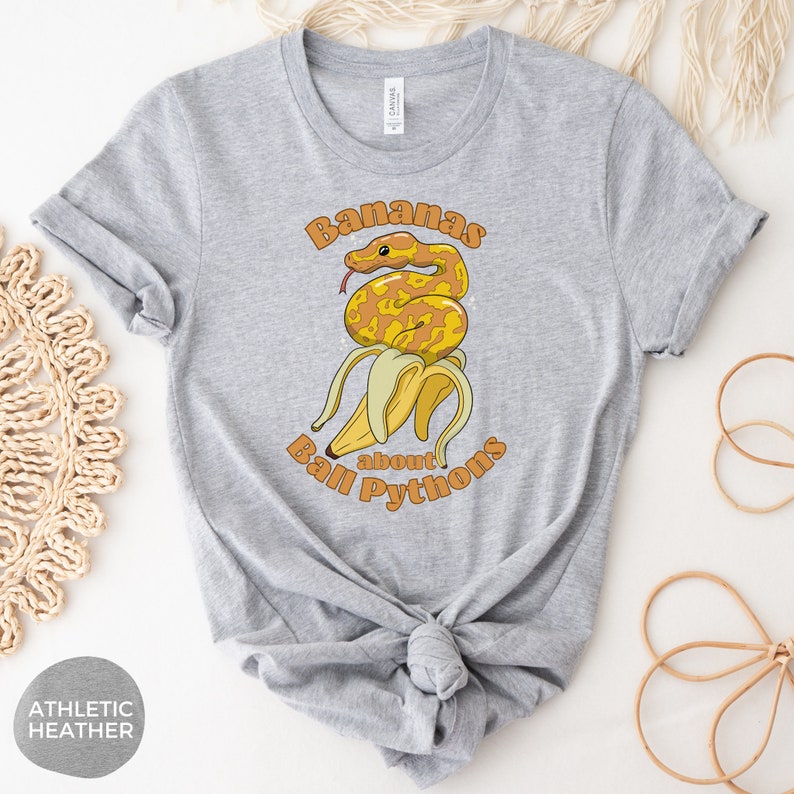 Op de afbeelding: Een grijs T-shirt met een afbeelding van een gele en oranje ball python die om een ​​bos bananen is gewikkeld. De tekst "Bananas about Ball Pythons" is in oranje en geel gedrukt.