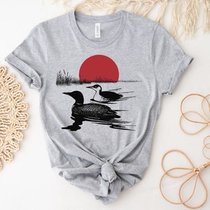 May include: Gray t-shirt with a black and white illustration of two loons swimming in a lake with a red sun setting in the background.
