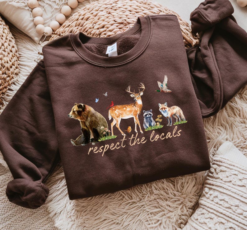 Op de afbeelding: Een bruine sweatshirt met een dierenprint met een beer, hert, wasbeer en vos. De tekst "respect the locals" staat eronder. De sweatshirt heeft een ronde hals en lange mouwen.