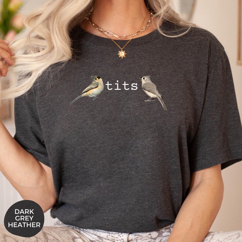 Puede incluir: Camiseta gris oscuro jaspeado con dos ilustraciones de p&aacute;jaros y la palabra "tits" en blanco. La camiseta tiene cuello redondo y mangas cortas. Una etiqueta circular en la esquina inferior izquierda dice "DARK GREY HEATHER".