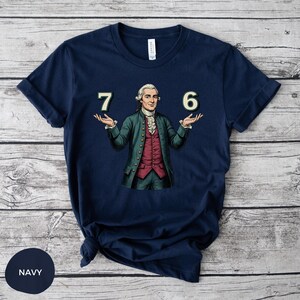 May include: Navy blue t-shirt featuring a historical figure with the numbers "7" and "6" on either side. The figure is dressed in a green coat, red vest, and white cravat. The shirt is made of a soft material.