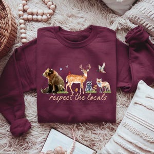 Op de afbeelding: Bordeaux sweatshirt met een aquarel illustratie van een beer, hert, wasbeer en vos, met de tekst "respect the locals". Het ontwerp bevat vogels en bloemen. De sweatshirt is gemaakt van een zacht materiaal.