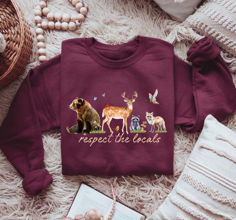 Op de afbeelding: Bordeaux sweatshirt met een aquarel illustratie van een beer, hert, wasbeer en vos, met de tekst "respect the locals". Het ontwerp bevat vogels en bloemen. De sweatshirt is gemaakt van een zacht materiaal.