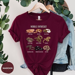 Puede incluir: Camiseta granate con la frase "NOODLE ENTHUSIAST" e ilustraciones de varias especies de serpientes, como la serpiente del ma&iacute;z, la boa de arena y la pit&oacute;n bola. La camiseta tambi&eacute;n muestra la culebra de liga, la pit&oacute;n alfombra, la serpiente de nariz de cerdo, la serpiente rey de California, la serpiente rey negra mexicana y la boa rosada.