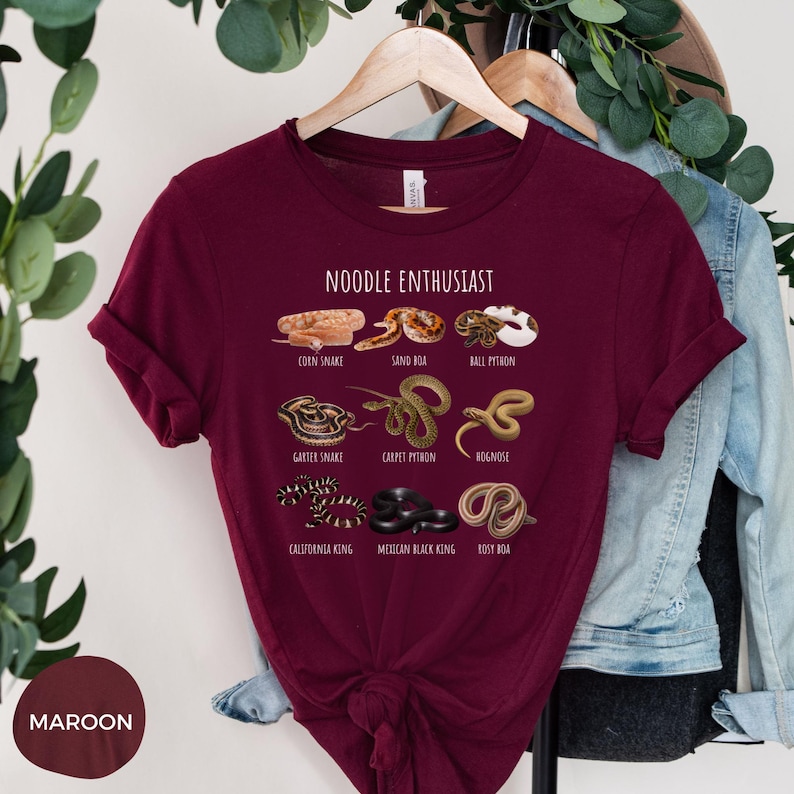 Puede incluir: Camiseta granate con la frase "NOODLE ENTHUSIAST" e ilustraciones de varias especies de serpientes, como la serpiente del ma&iacute;z, la boa de arena y la pit&oacute;n bola. La camiseta tambi&eacute;n muestra la culebra de liga, la pit&oacute;n alfombra, la serpiente de nariz de cerdo, la serpiente rey de California, la serpiente rey negra mexicana y la boa rosada.