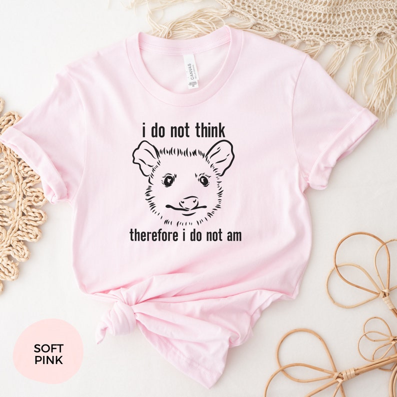 May include: A soft pink t-shirt featuring a black and white illustration of an opossum and the text "i do not think therefore i do not am"