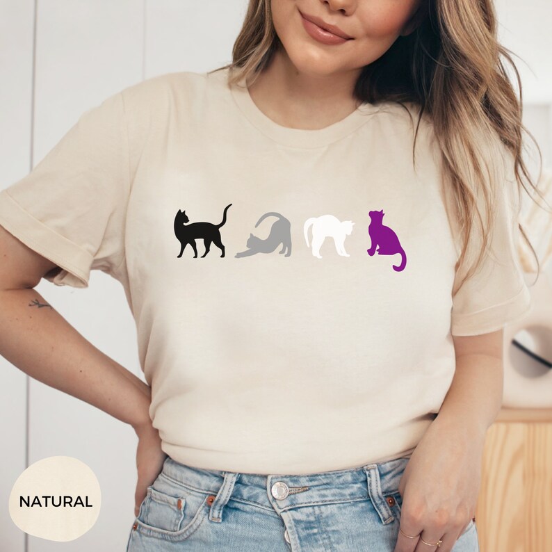 May include: A beige t-shirt with a black, gray, and purple cat silhouette design. The cats are arranged in a line, representing the asexual pride flag.