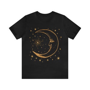 Pode incluir: Camiseta preta com um design de lua crescente e estrelas douradas. A lua tem um rosto e um brilho de estrelas atr&aacute;s.