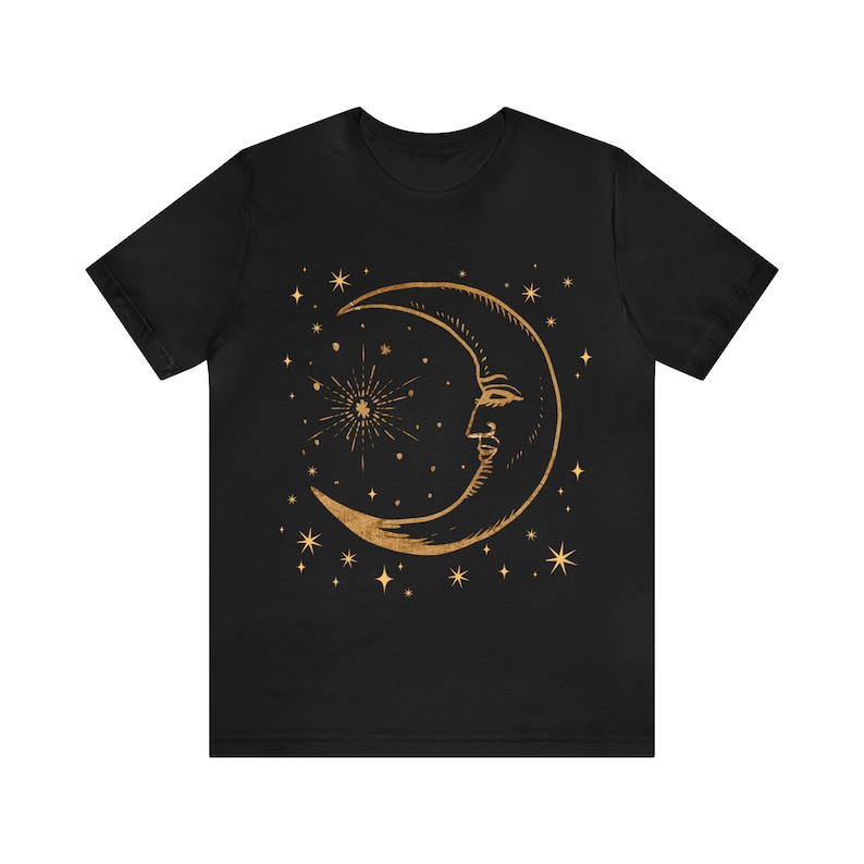 Pode incluir: Camiseta preta com um design de lua crescente e estrelas douradas. A lua tem um rosto e um brilho de estrelas atr&aacute;s.