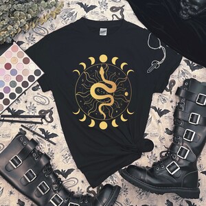 May include: Black t-shirt with a gold snake and moon design. The snake is coiled around a sun with a starburst pattern. The moon phases are around the sun and snake.