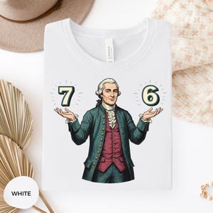 May include: White t-shirt featuring a cartoon illustration of a man in a green coat and red vest, holding up the numbers 7 and 6. The shirt has a crew neck and the word "WHITE" is printed on a white circle.