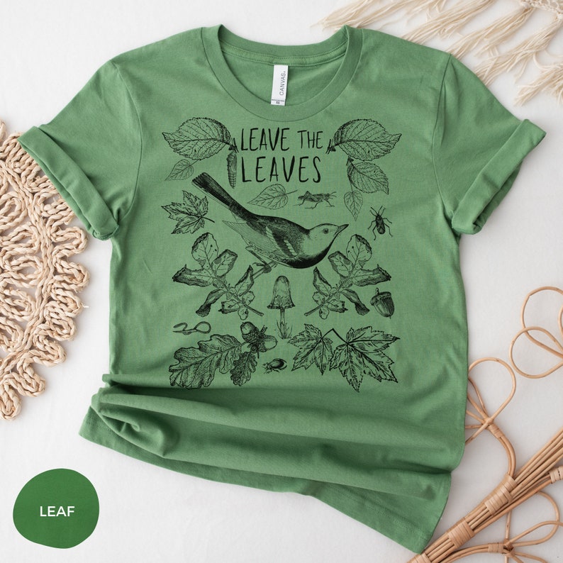 May include: Green t-shirt with a black and white illustration of a bird, leaves, and other nature elements. The text "Leave the Leaves" is printed on the shirt.