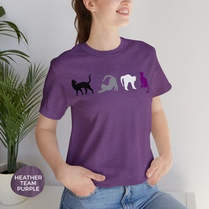 May include: A heather purple t-shirt with a row of four black, gray, white, and purple cat silhouettes. The cats are arranged in a line, with the black cat on the left and the purple cat on the right. The shirt has a crew neck and short sleeves.