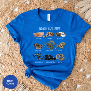 Puede incluir: Una camiseta azul real con la frase "NOODLE ENTHUSIAST" y dibujos de varias especies de serpientes, como la serpiente del ma&iacute;z y la pit&oacute;n bola. La camiseta tambi&eacute;n tiene el texto "TRUE ROYAL" en la parte inferior izquierda.