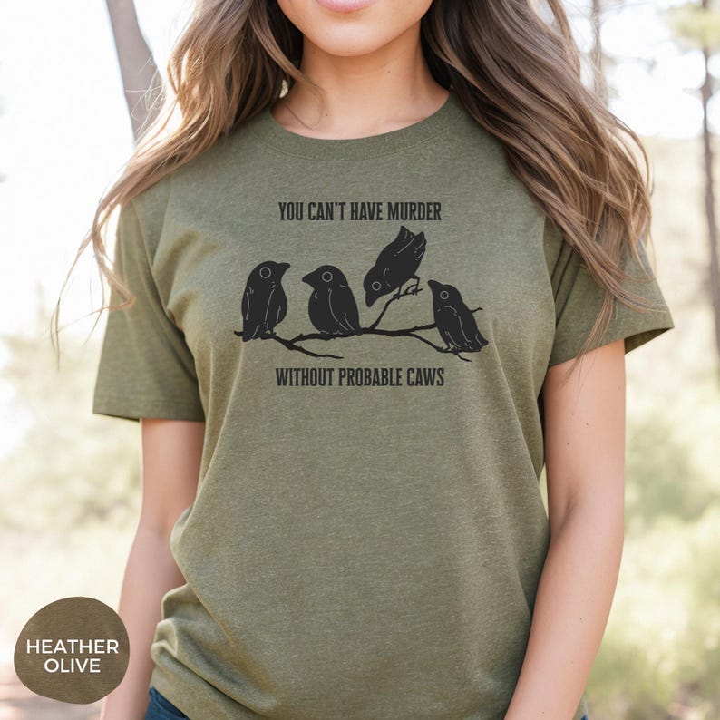 May include: Heather olive green t-shirt with the text "YOU CAN'T HAVE MURDER WITHOUT PROBABLE CAWS" and a graphic of four black birds perched on a branch. The shirt is a short-sleeved crew neck.
