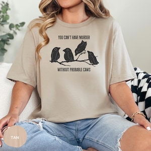 May include: Tan-colored t-shirt featuring a graphic of four black birds perched on a branch. The text "YOU CAN'T HAVE MURDER WITHOUT PROBABLE CAWS" is printed above and below the birds. The shirt is made of soft material.