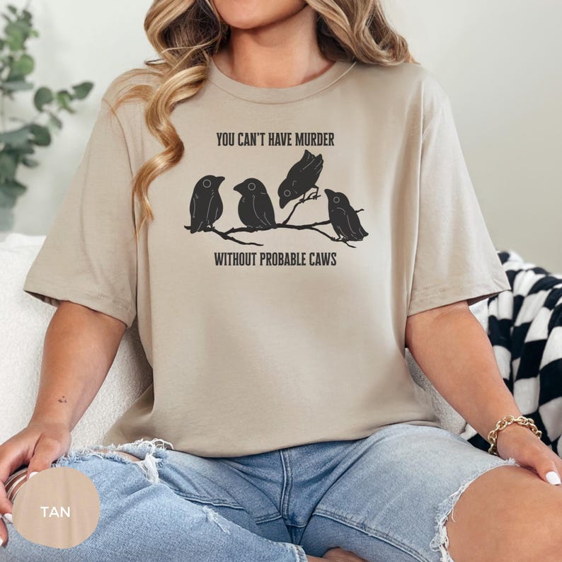May include: Tan-colored t-shirt featuring a graphic of four black birds perched on a branch. The text "YOU CAN'T HAVE MURDER WITHOUT PROBABLE CAWS" is printed above and below the birds. The shirt is made of soft material.