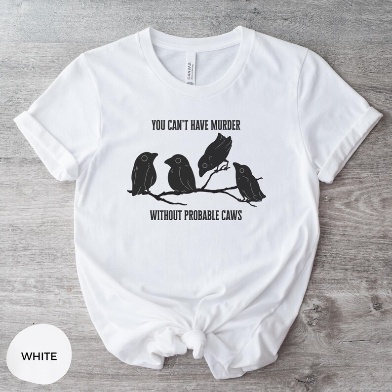May include: White t-shirt with a crew neck and rolled-up sleeves. The shirt features a black graphic of four birds perched on a branch, with the text "YOU CAN'T HAVE MURDER WITHOUT PROBABLE CAWS."