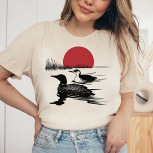May include: A white t-shirt with a black and white illustration of two loons swimming in a lake with a red sun setting in the background.