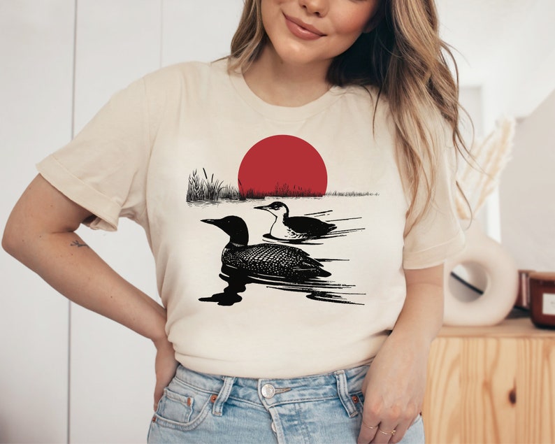 May include: A white t-shirt with a black and white illustration of two loons swimming in a lake with a red sun setting in the background.