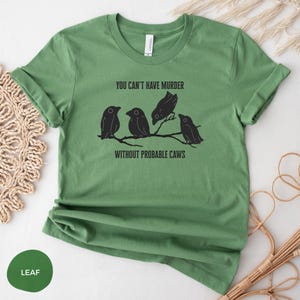 May include: A leaf green t-shirt with the text "YOU CAN'T HAVE MURDER WITHOUT PROBABLE CAWS" and a graphic of four black birds perched on a branch. The shirt has rolled-up sleeves.