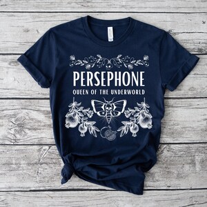 May include: A navy blue t-shirt with a white graphic design featuring the text "PERSEPHONE QUEEN OF THE UNDERWORLD" with a stylized moth and pomegranate imagery.