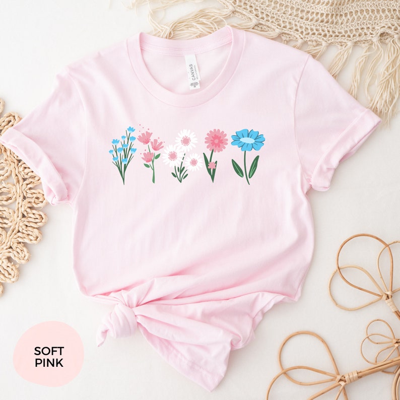 May include: A soft pink t-shirt with a floral design featuring blue, pink, and white flowers. The flowers are arranged in a row across the front of the shirt.