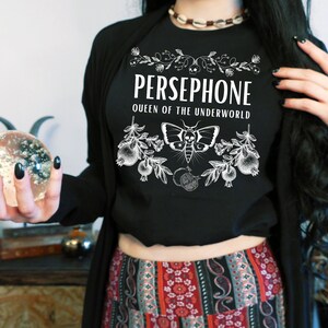May include: Black t-shirt with white text that reads "PERSEPHONE QUEEN OF THE UNDERWORLD" and a graphic of a moth and pomegranate branches.