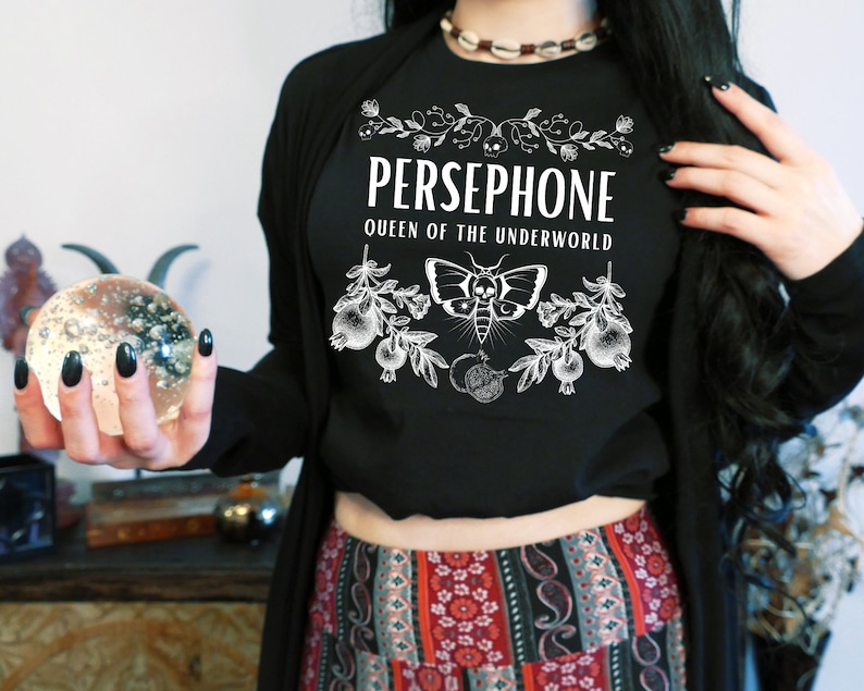 May include: Black t-shirt with white text that reads "PERSEPHONE QUEEN OF THE UNDERWORLD" and a graphic of a moth and pomegranate branches.