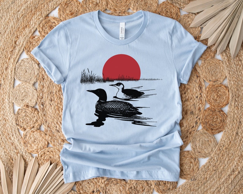 May include: A light blue t-shirt with a black and white illustration of two loons swimming in a lake with a red sun setting in the background.