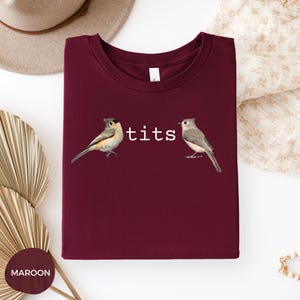 Puede incluir: Camiseta granate con la palabra "tits" en blanco y dos ilustraciones de p&aacute;jaros. Las aves est&aacute;n representadas con plumas grises, marrones y blancas. La camiseta est&aacute; doblada, con una etiqueta visible en el cuello.