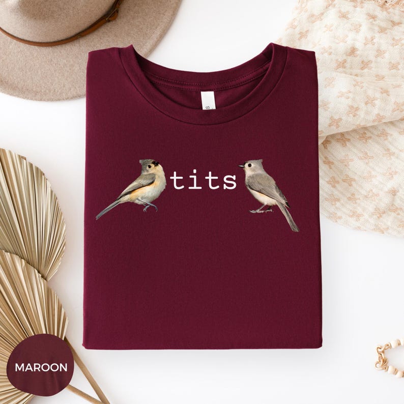Puede incluir: Camiseta granate con la palabra "tits" en blanco y dos ilustraciones de p&aacute;jaros. Las aves est&aacute;n representadas con plumas grises, marrones y blancas. La camiseta est&aacute; doblada, con una etiqueta visible en el cuello.