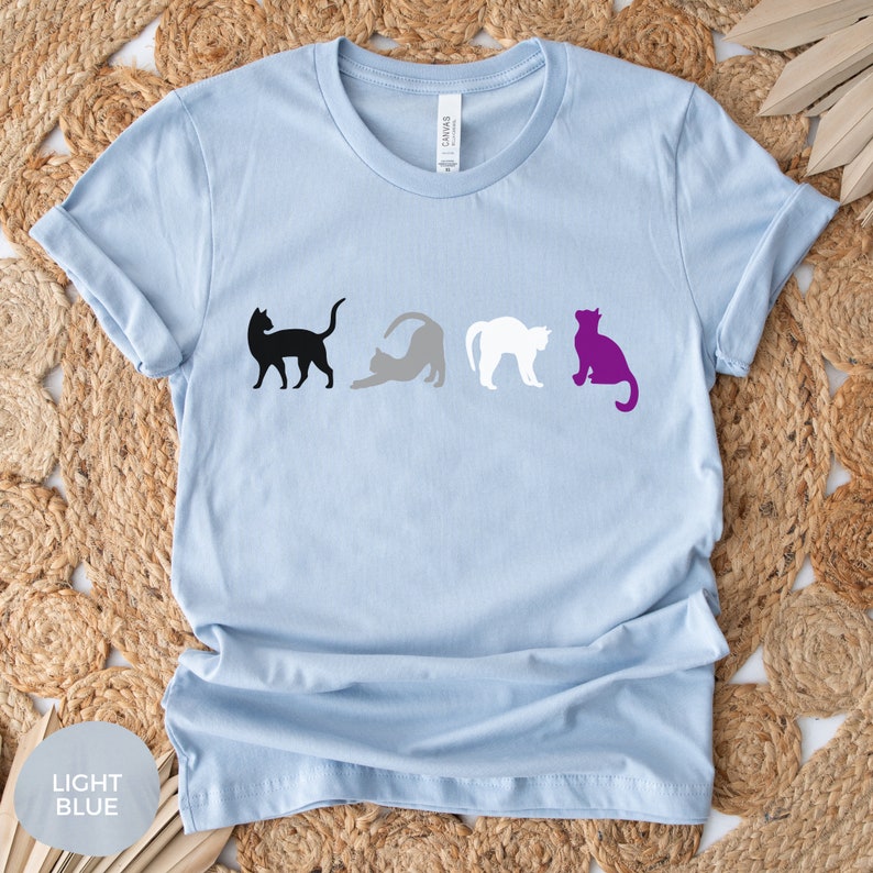 May include: Light blue t-shirt with a black, gray, white, and purple cat silhouette design. The cats are arranged in a row, representing the asexual pride flag.