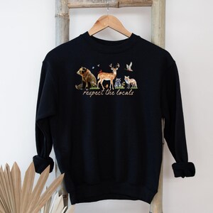 Op de afbeelding: Zwart sweatshirt met een aquarel illustratie van een beer, hert, wasbeer en vos. De tekst "respect the locals" is onder de dieren gedrukt. Het sweatshirt hangt aan een houten hanger.